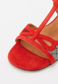 Sandalo rosso in suede con cinturini incrociati e pannello geometrico a motivi bianchi e neri sul tallone. La texture in suede aggiunge morbidezza.