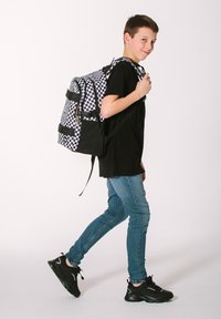 Zaino a scacchi bianco e nero con dettagli neri, spallacci imbottiti e tasche laterali. Indossato da una persona in una maglietta nera e jeans blu.