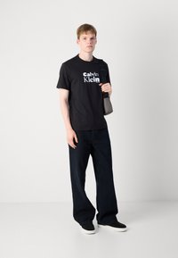 Juodas medvilninis marškinėliai su balta "Calvin Klein" logo, derinami su tamsiai mėlynomis plačiomis džinsomis ir juoda avalyne. Minimalistinis dizainas, kasdienis stilius.