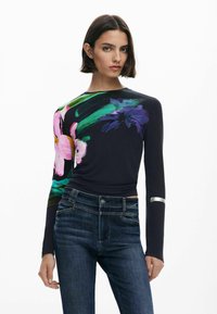 Haut à manches longues noir avec un imprimé floral vibrant en rose, violet et vert. Dispose d'un accent métallique sur une manche. Associé à un jean bleu.