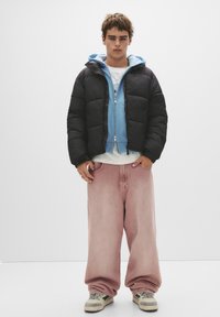 Sort pufferjakke, lyseblå hoodie, hvid t-shirt og lyserøde baggy jeans. Grå og creme sneakers med detaljer. Ensfarvet baggrund.