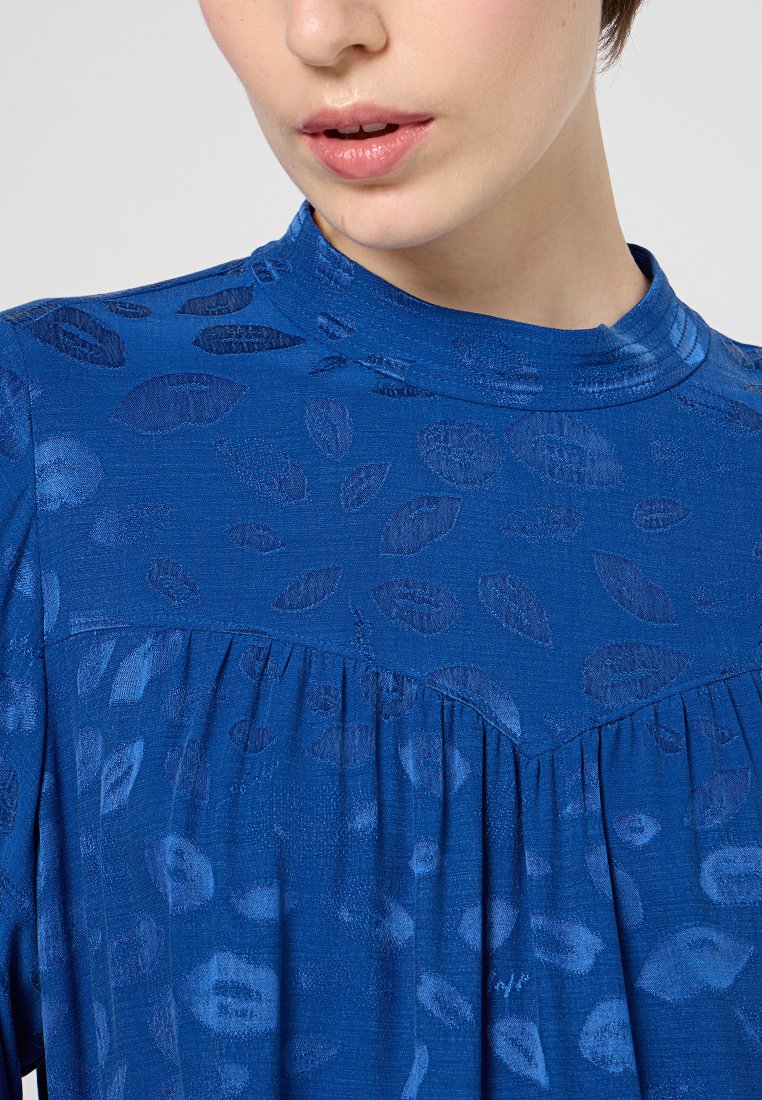 Blaue Bluse mit einem strukturierten Blattmuster, hohem Kragen und gefaltetem Design an der Brust. Glattes Material mit einem leichten Glanz.