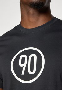 T-shirt en coton noir avec un grand motif graphique circulaire blanc représentant le numéro "90" au centre, manches courtes et col rond.