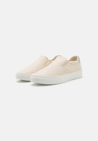 Beige slip-on sko med teksturert stoffoverdel, hvit gummisåle og elastiske sidepaneler for enkel bruk. Diskret logodetalj på hælen.