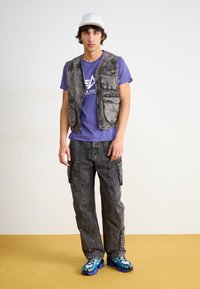 Vest din denim gri cu multiple buzunare purtat peste un tricou violet, asociat cu pantaloni cargo gri și adidași cu model albastru. Căciulă albă pe cap.