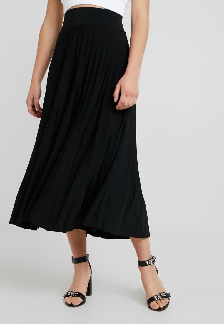 Femme portant une jupe midi plissée noire, un crop top blanc et des sandales à talons hauts noires avec des brides à la cheville sur un fond gris clair.