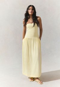 Abito lungo giallo senza spalline con corpetto aderente e gonna arricciata. Realizzato in tessuto morbido, presenta tasche laterali e una silhouette fluida.