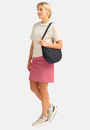 Donna sorridente, in piedi con camicia beige chiaro, gonna rosa, sneakers beige e una borsa a tracolla nera con disegno di impronta di zampa.
