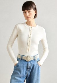 Cremfarbener, gerippter Langarm-Top mit Knopfleiste, kombiniert mit lockeren blauen Denim-Jeans und einem gewebten Gürtel mit runder Schnalle.