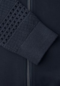 Dunkelblauer Strickpulloverärmel mit strukturierten Mustern, der auf einem passenden marineblauen Stoffhintergrund mit einem vertikalen silbernen Reißverschluss ruht.