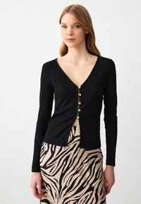 Zwarte geribbelde top met lange mouwen en een diepe V-hals, voorzien van een rij gouden knopen aan de voorkant, gecombineerd met een beige rok met zwarte zebrapatronen.