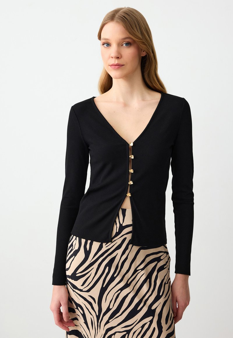 Zwarte geribbelde top met lange mouwen en een diepe V-hals, voorzien van een rij gouden knopen aan de voorkant, gecombineerd met een beige rok met zwarte zebrapatronen.