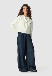 Veste blanche ajustée avec des boutons dorés et deux poches poitrine, associée à un pantalon en denim noir à jambes larges. Tissu lisse, design décontracté.