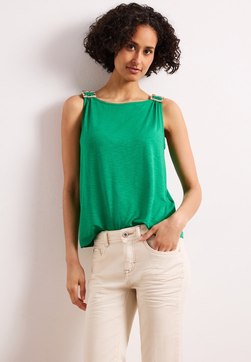 Femme aux cheveux bouclés portant un haut sans manches vert avec des bretelles à boucle et un pantalon clair, debout contre un mur blanc.