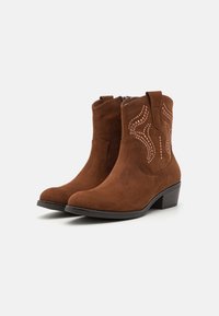 Bottines en daim marron avec des coutures décoratives et des accents métalliques, dotées d'un petit talon et d'une fermeture éclair sur le côté pour un enfilage facile.