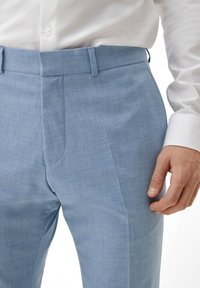 Helle blaue maßgeschneiderte Hosen aus einem strukturierten Stoff. Sie verfügen über eine flache Vorderseite, Gürtelschlaufen und seitliche Taschen. Kombiniert mit einem weißen Hemd.