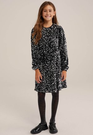 Svart klänning täckt av reflektiva paljetter, lång ärm med rynkade manschetter, knälång nederkant. Bärs med mörka, texturerade strumpbyxor och blanka svarta skor.