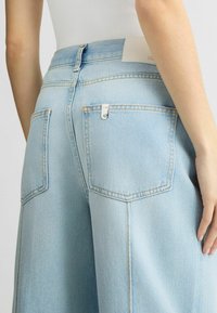 Jeans de mezclilla azul claro con cintura alta, dos bolsillos traseros y costuras amarillas. Presentan un parche de logo blanco en la cinturilla.