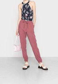 Top halter floral en azul marino, combinado con joggers sueltos de color rosa empolvado. Sandalias blancas con suelas negras, sosteniendo un pequeño bolso rosa.