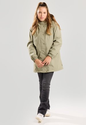 Ragazza giovane in piedi indossa una giacca verde con bottoni, jeans scuri e sneakers bianchi su uno sfondo chiaro e uniforme.