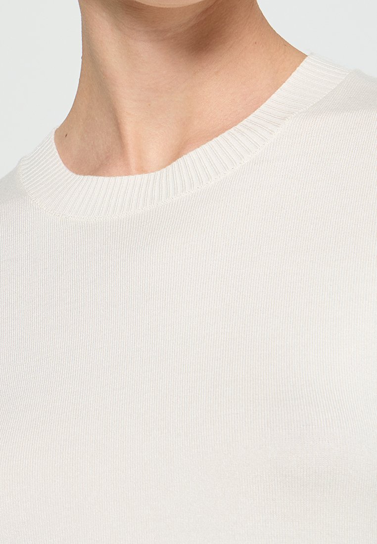 Gros plan sur une personne portant un pull à col rond blanc en côte, montrant l'encolure et la partie supérieure de la poitrine.