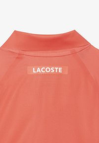 Korallfärgad polo med mjuk textur och en kontrasterande vit "LACOSTE" logotyp på baksidan av kragen, med en modern och sportig design.