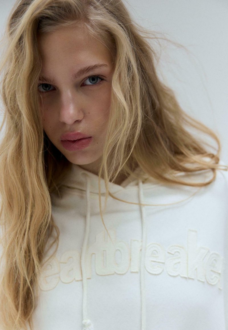 Sweat à capuche blanc avec le mot "heartbreaker" en lettres en relief ton sur ton. Texture douce, cordons de serrage au niveau du col, et cheveux blonds clairs ondulés au premier plan.