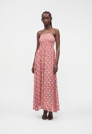 Femme portant une robe maxi sans manches à motifs roses et rouges avec de fines bretelles, debout sur un fond blanc uni.