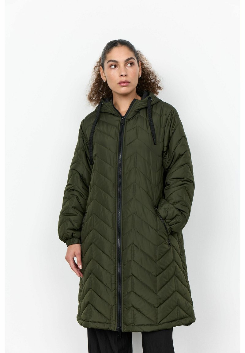Femme aux cheveux bouclés portant une longue veste matelassée vert olive avec une capuche, une fermeture éclair noire et des cordons noirs, debout sur un fond blanc.