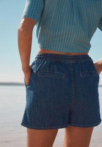 Pantaloni scurți din denim cu talie elastică, două buzunare în spate și croială lejeră. Bluza cu mâneci scurte, de culoare albastru deschis, este vizibilă deasupra taliei.