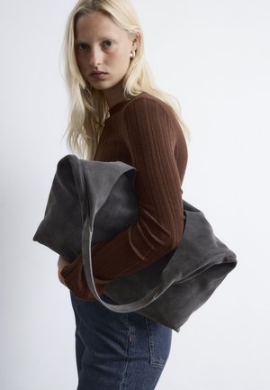 Gray suede tote bag with soft, rounded edges, featuring a single strap, held close to the body against a brown ribbed long-sleeve top.