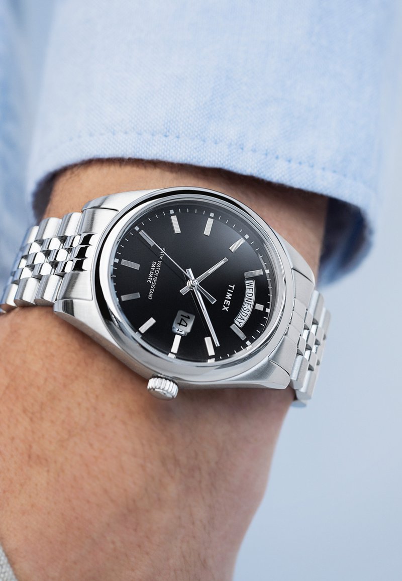 Montre en acier inoxydable argenté avec un cadran noir, des index horaires argentés et un affichage de la date. Elle possède un bracelet à maillons et un boîtier rond.
