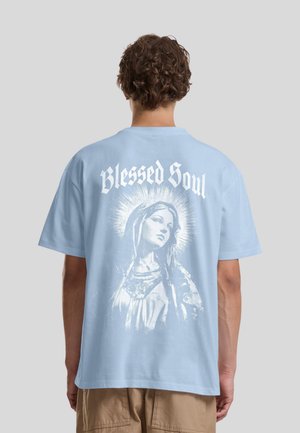 Personne portant une chemise bleu clair avec un graphisme blanc d'une femme avec une auréole et le texte "Blessed Soul" dans le dos.