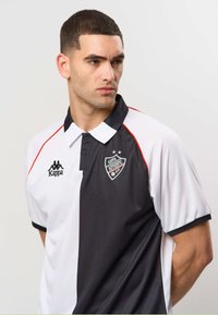 Jeune homme aux cheveux courts portant un maillot de sport Kappa noir et blanc avec des logos, regardant vers sa gauche sur un fond uni.