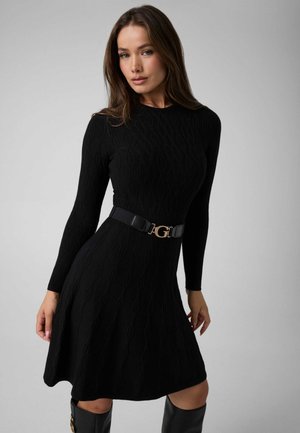 Femme portant une robe en maille noire ajustée à manches longues avec un motif texturé et une ceinture noire, debout devant un fond uni.