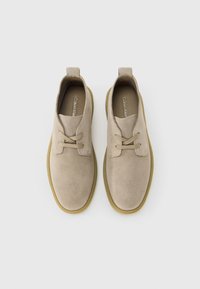 Pantofi din suede bej cu șireturi și tălpi de culoare maro deschis, văzuți dintr-o parte de sus pe un fundal alb simplu.