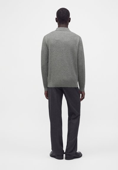Homme debout de dos, portant un pull gris à manches longues, un pantalon gris foncé et des chaussures noires sur fond blanc.