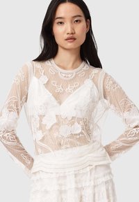 Durchsichtige weiße Langarmbluse mit floraler Stickerei, strukturiertem Overlay, runder Ausschnitt und Spitzenakzenten am Saum.