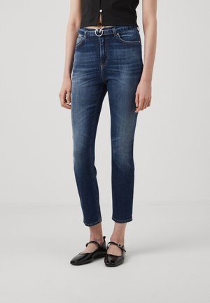 Jeans in denim blu con una vestibilità slim, vita alta e passanti per cintura, caratterizzati da una fibbia circolare argentata. Abbinati a ballerine nere.
