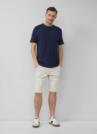 Marineblauw T-shirt met korte mouwen gecombineerd met lichtbeige shorts. De schoenen zijn wit en beige sneakers. Het model staat met de handen in de zakken.
