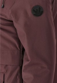 Rękaw bordowej kurtki z czarną okrągłą naszywką z logo Whistler Denmark, widocznym przeszyciem szwu i patką kieszeni.