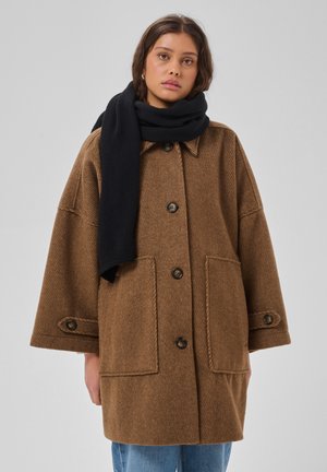 Jonge vrouw die een oversized bruine jas draagt met zwarte knopen en grote zakken, gecombineerd met een zwarte sjaal en blauwe spijkerbroek.