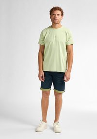 Camiseta de algodón de color verde claro con cuello redondo, combinada con pantalones cortos de mezclilla oscura con dobladillos enrollados. Zapatillas blancas completan el conjunto.