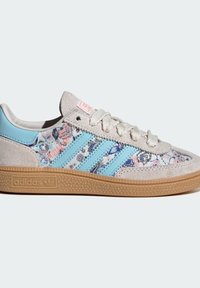 Baskets basses Adidas avec des rayures bleu clair, daim beige, panneaux en tissu fleuri, lacets blancs et semelle en gomme embossée du logo Adidas.