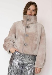 Nepbont cropped jack in lichtbeige met een hoge kraag, voorzien van vier knopen en contrasterende grijze stiksels. Model draagt zilverkleurige broek met slangenprint.