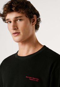 Črna bombažna majica z okrolim ovratnikom, s rdečim izvezenim napisom na prsih: "Rojen za zvezdo rocka, prisiljen delati."