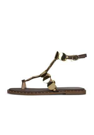 Sandalia plana marrón con tira en el tobillo y tachuelas decorativas circulares doradas a lo largo de las tiras y el borde de la suela.