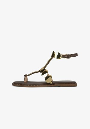 Bruine platte sandaal met enkelbandje en gouden ronde decoratieve studs langs de bandjes en de rand van de zool.