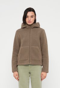 Καφέ μάλλινο μπουφάν με κουκούλα, μπροστινό φερμουάρ και δύο μεγάλες τσέπες. Το υλικό είναι υφή με απαλό, ζεστό ύφος.