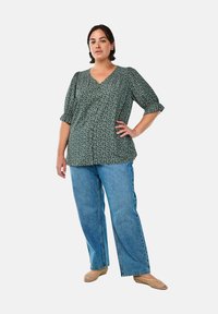Blusa verde con fantasia floreale, collo a V e maniche a sbuffo, abbinata a jeans a gamba larga di un blu chiaro e ballerine beige intrecciate.